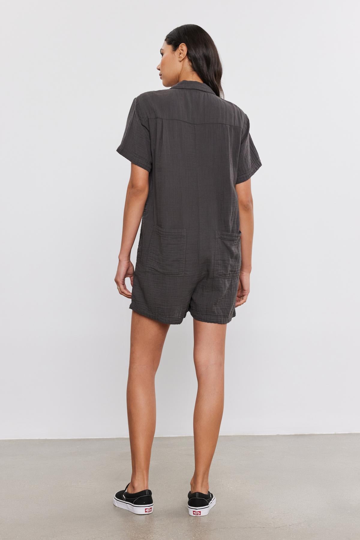   A woman stands facing away from the camera, wearing a gray DANE COTTON GAUZE romper from Velvet by Graham & Spencer and black sneakers with white soles. 