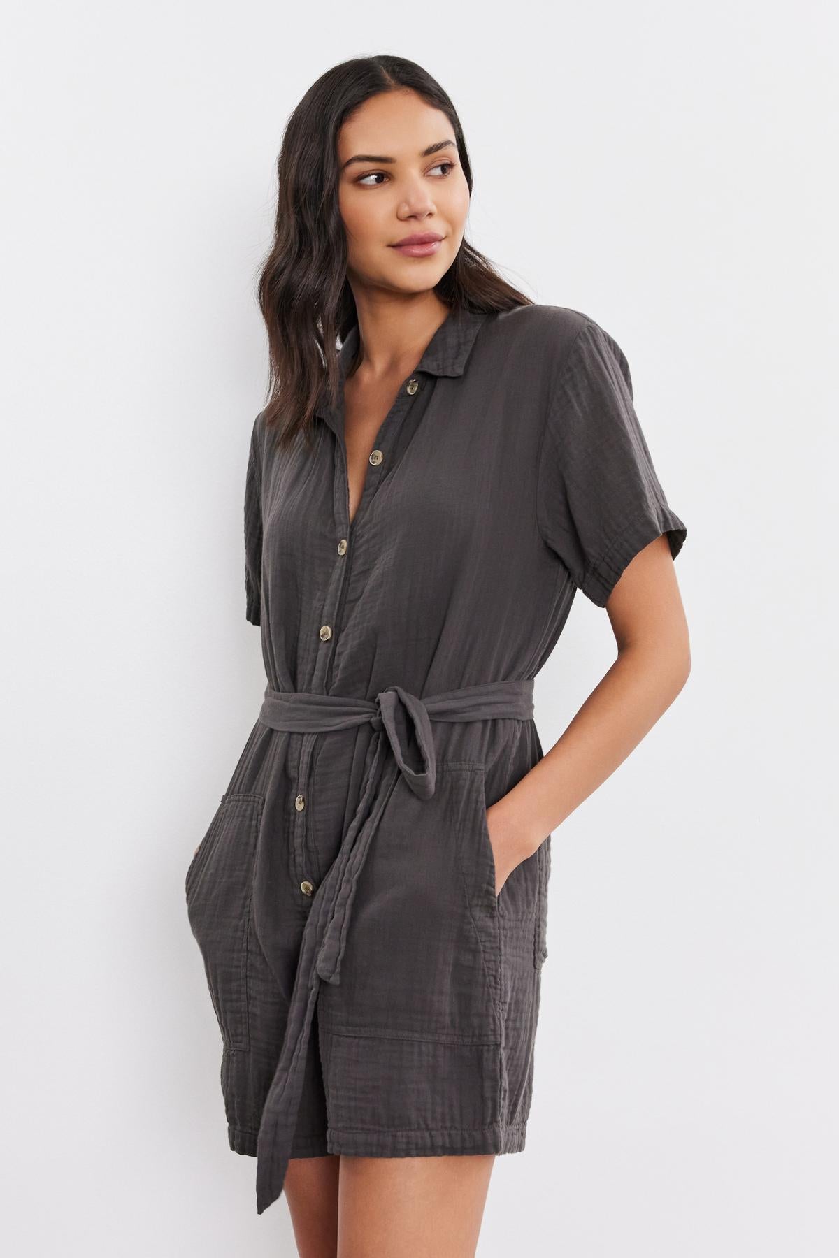   A woman models a casual grey button front DANE COTTON GAUZE ROMPER with short sleeves, a collar, and a tied waist. She looks to the side with a slight smile. (Brand Name: Velvet by Graham & Spencer) 