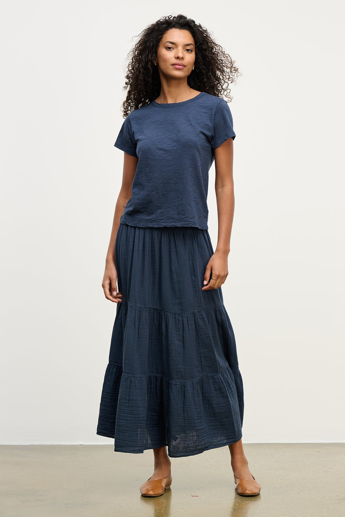   A person wearing a navy T-shirt and the DANIELLE COTTON GAUZE SKIRT by Velvet by Graham & Spencer, featuring a flowing midi-to-maxi length with an elastic waist, stands gracefully against a plain background. 