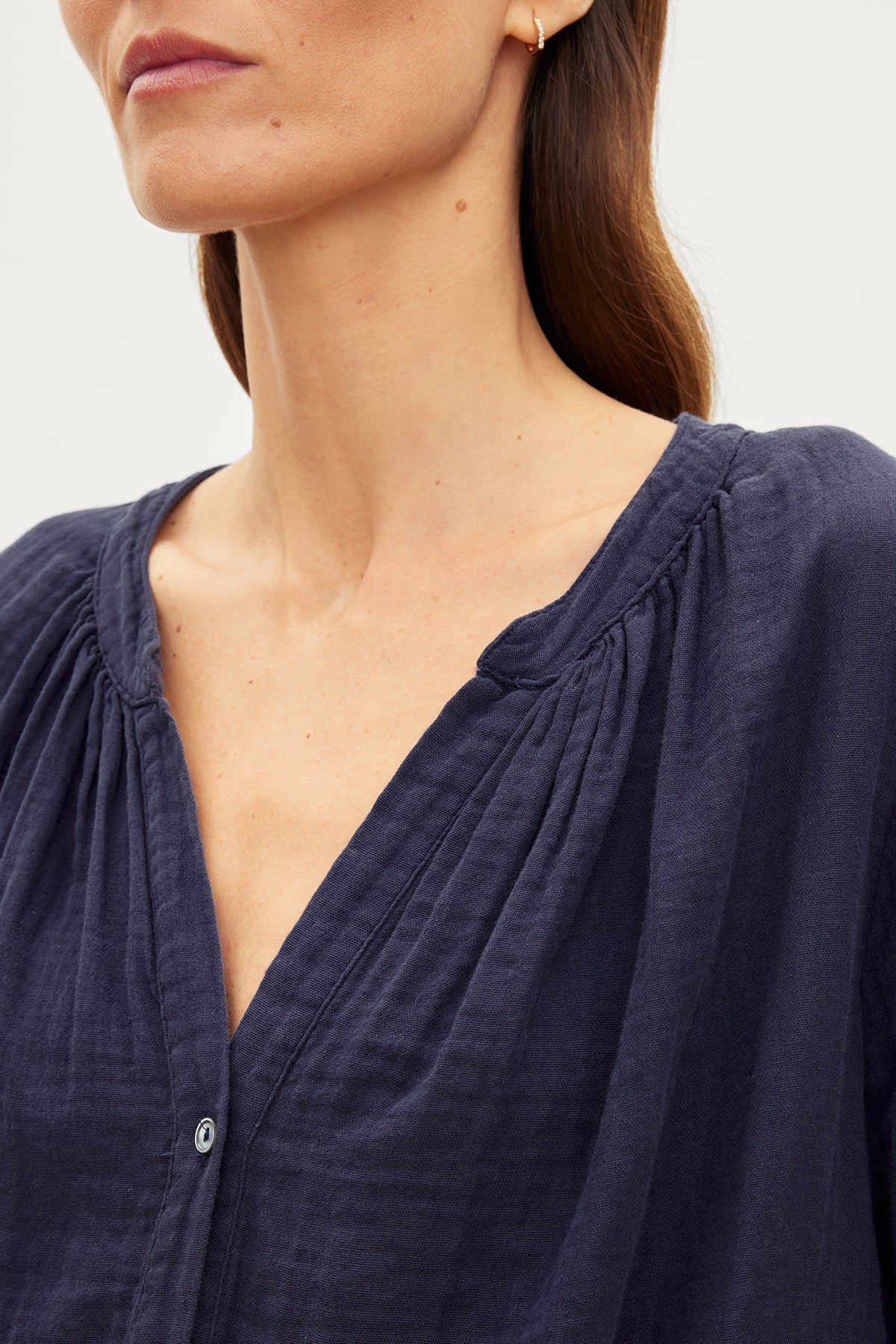 Close-up of a woman wearing the DEANN COTTON GAUZE TOP by Velvet by Graham & Spencer, which is a dark blue, cotton gauze top with a gathered v-neckline and button front. The image shows her from the neck to the mid-chest level, highlighting the texture and design of the blouse.-35955697549505
