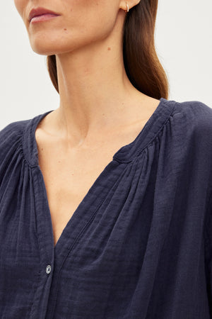 Close-up of a woman wearing the DEANN COTTON GAUZE TOP by Velvet by Graham & Spencer, which is a dark blue, cotton gauze top with a gathered v-neckline and button front. The image shows her from the neck to the mid-chest level, highlighting the texture and design of the blouse.