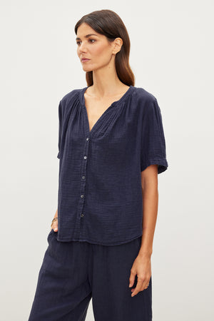 A person stands against a plain background wearing a dark blue, short-sleeved, button-front DEANN COTTON GAUZE TOP by Velvet by Graham & Spencer with a matching pair of pants.