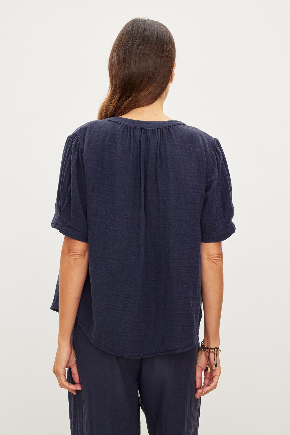 A person with long hair is seen from behind, wearing a DEANN COTTON GAUZE TOP by Velvet by Graham & Spencer with a button front and matching blue pants. The background is plain and light-colored.-35955697483969