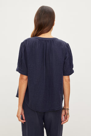 A person with long hair is seen from behind, wearing a DEANN COTTON GAUZE TOP by Velvet by Graham & Spencer with a button front and matching blue pants. The background is plain and light-colored.