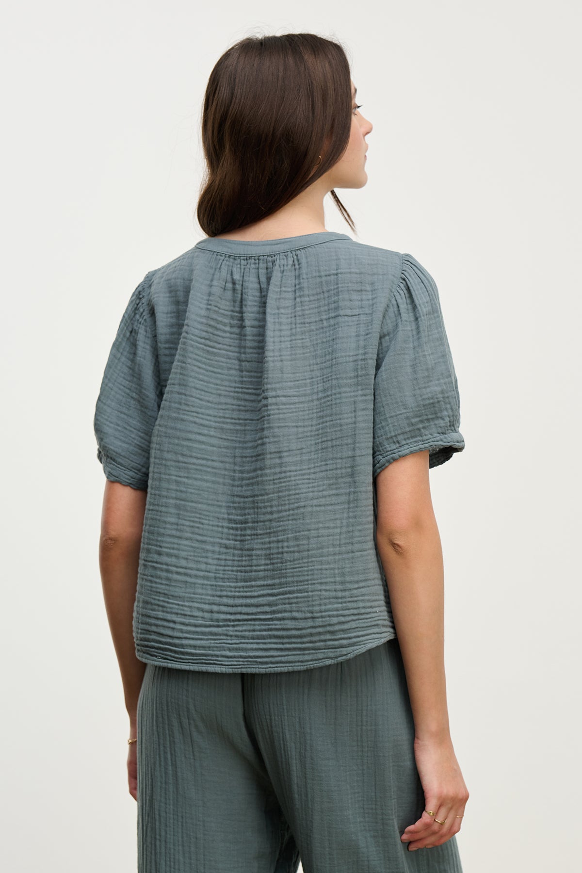 A person is seen from behind wearing the DEANN COTTON GAUZE TOP by Velvet by Graham & Spencer, featuring a textured, loose-fitting design in gray-green with a subtle v-neckline and matching pants, crafted from soft cotton gauze against a plain backdrop.-38662568313025