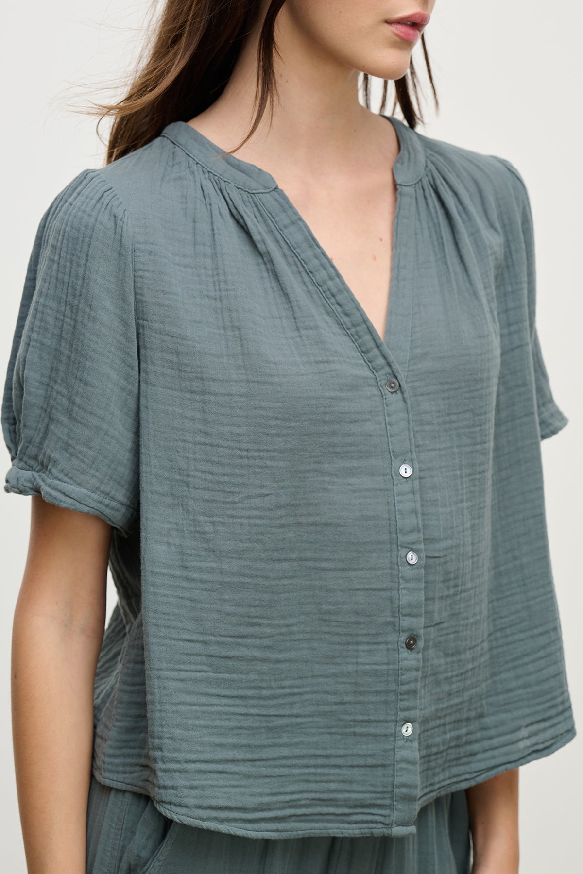 A person wears the DEANN COTTON GAUZE TOP by Velvet by Graham & Spencer, a grey, short-sleeved blouse with a button front. Made of soft cotton gauze, it features a delicate v-neckline, combining comfort and style effortlessly.-38662568345793