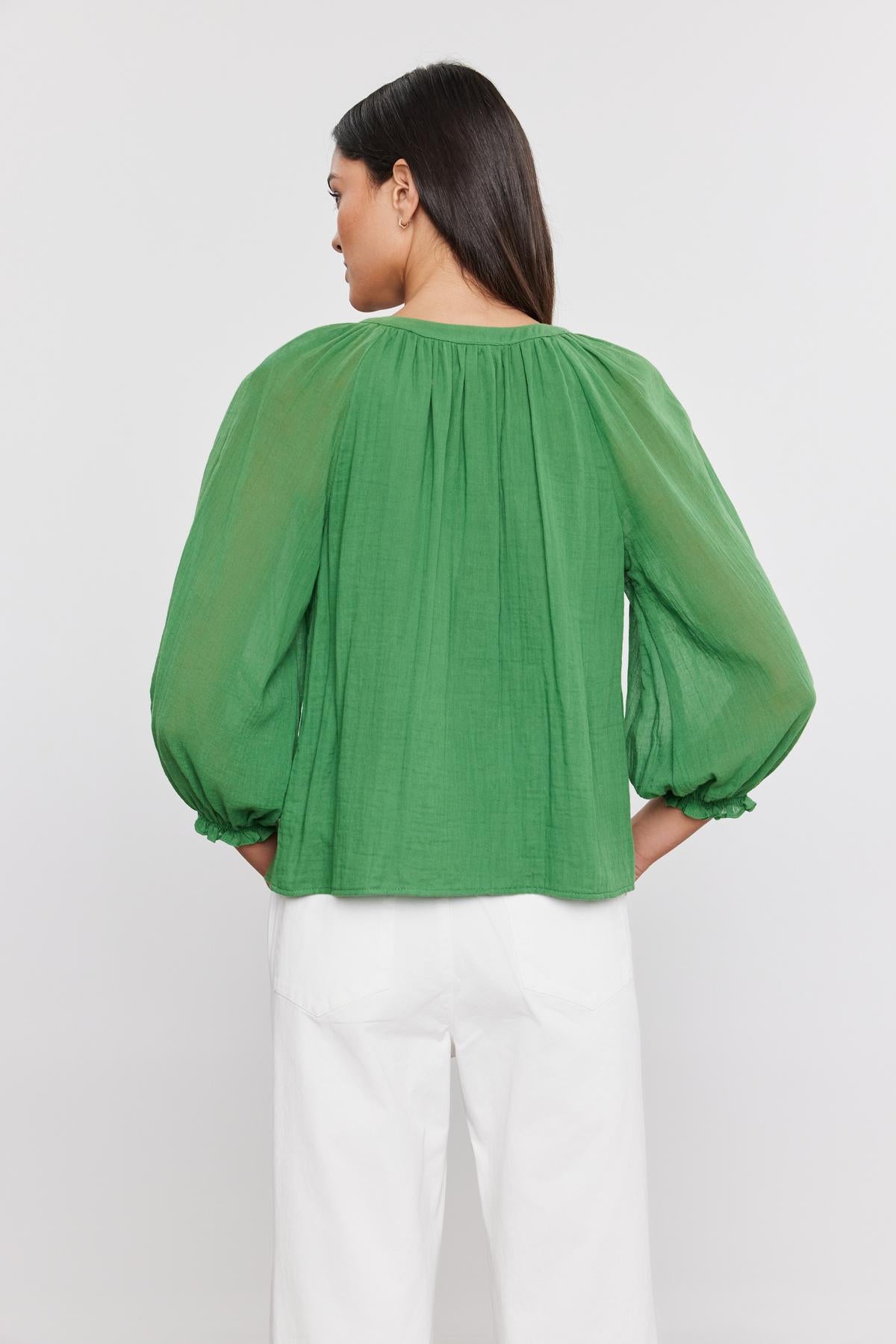   Rear view of a woman wearing a DENICE COTTON GAUZE TOP by Velvet by Graham & Spencer with sheer sleeves and white pants, standing against a plain background. 