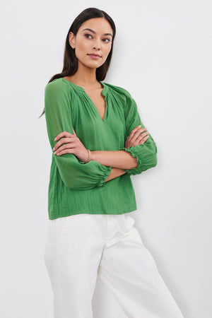 A woman stands with arms crossed, wearing a green DENICE COTTON GAUZE TOP by Velvet by Graham & Spencer and white pants, against a plain white background.