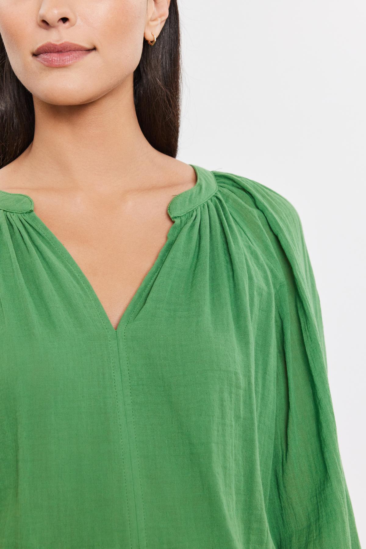   Close-up of a woman in a DENICE COTTON GAUZE TOP by Velvet by Graham & Spencer in a green blouse with a v-neckline and gathered details, featuring sheer sleeves, focusing on the garment and the lower half of her face. 