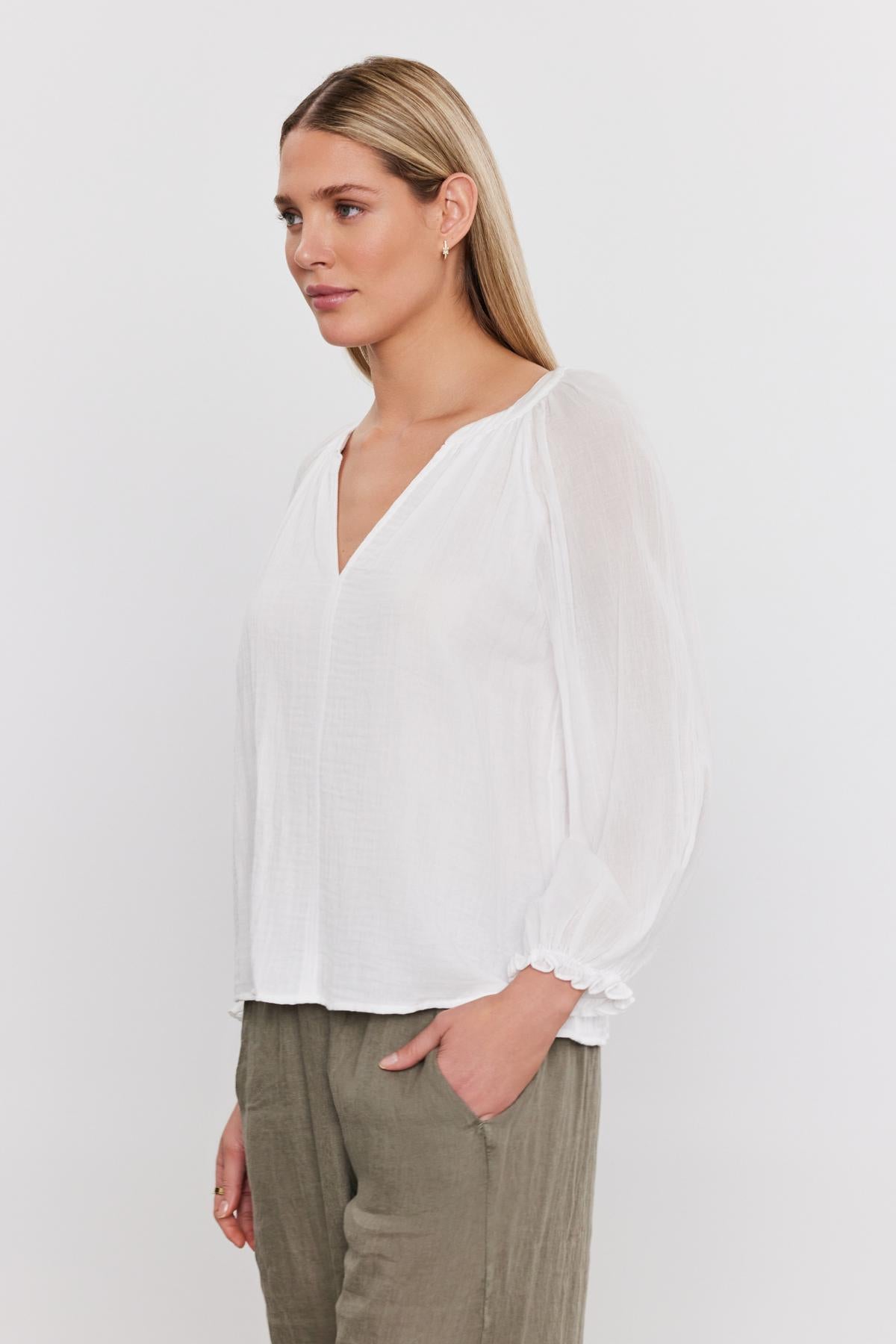   Profile view of a woman with blonde hair wearing a Denice Cotton Gauze Top from Velvet by Graham & Spencer, with sheer sleeves and green trousers, standing against a plain background. 