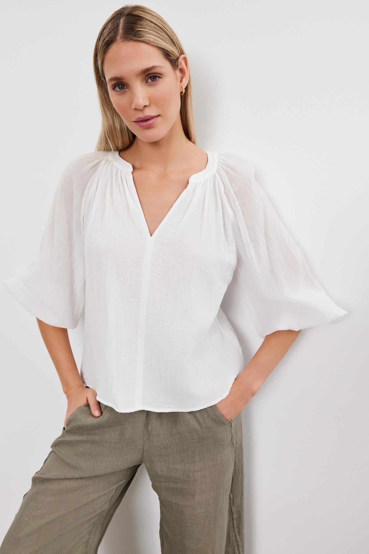   A woman in a Denice Cotton Gauze Top by Velvet by Graham & Spencer with sheer sleeves and green trousers stands against a plain background, looking directly at the camera. 