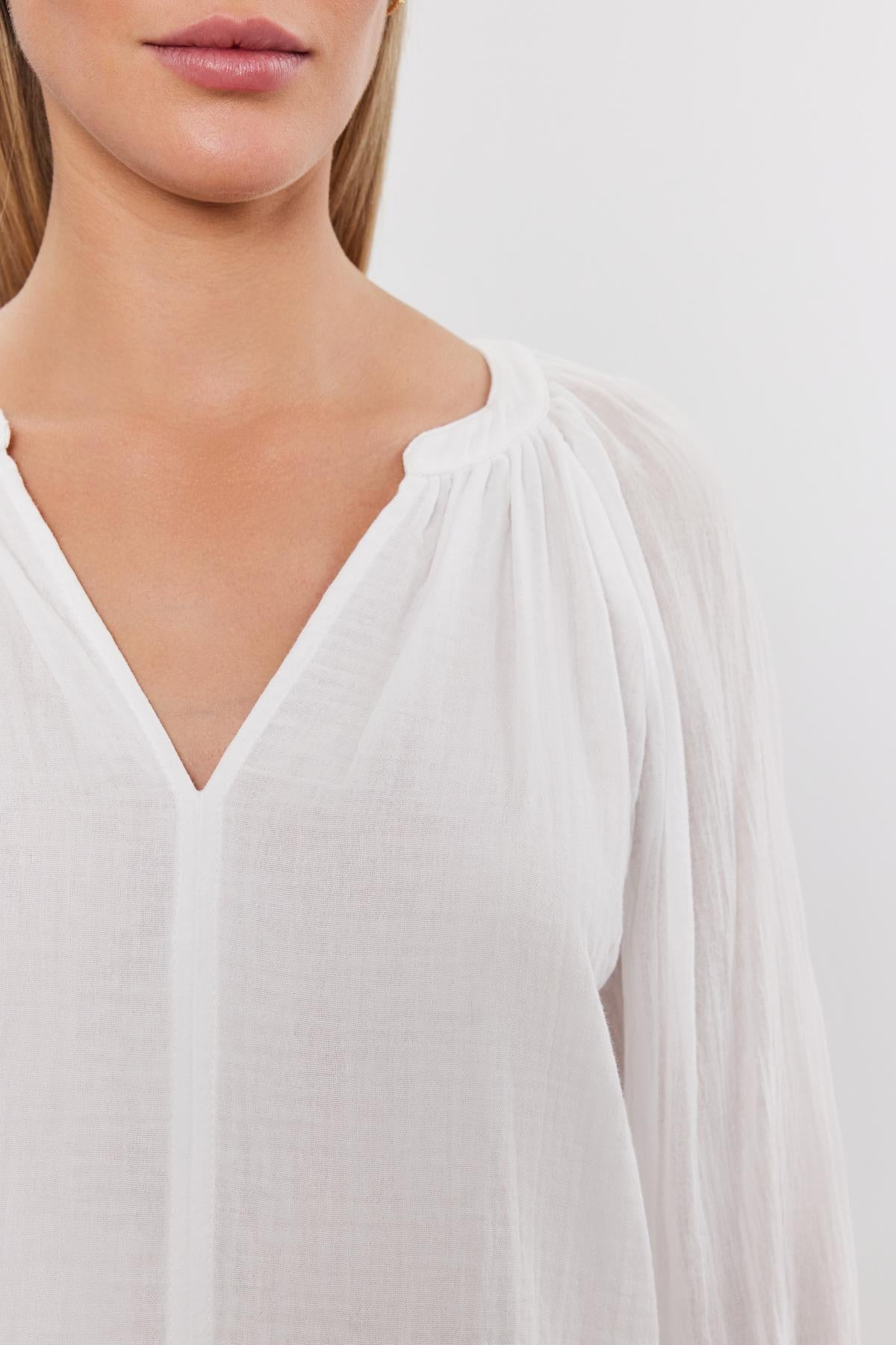   Close-up of a woman wearing a white DENICE COTTON GAUZE TOP by Velvet by Graham & Spencer with gathered detailing at the v-neckline. Only her neck and part of her chest are visible. 