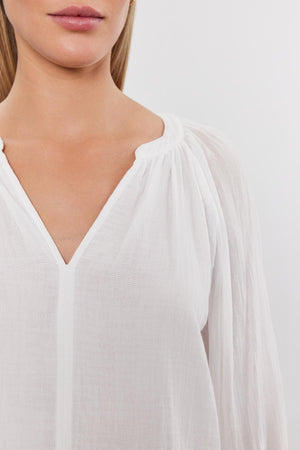 Close-up of a woman wearing a white DENICE COTTON GAUZE TOP by Velvet by Graham & Spencer with gathered detailing at the v-neckline. Only her neck and part of her chest are visible.