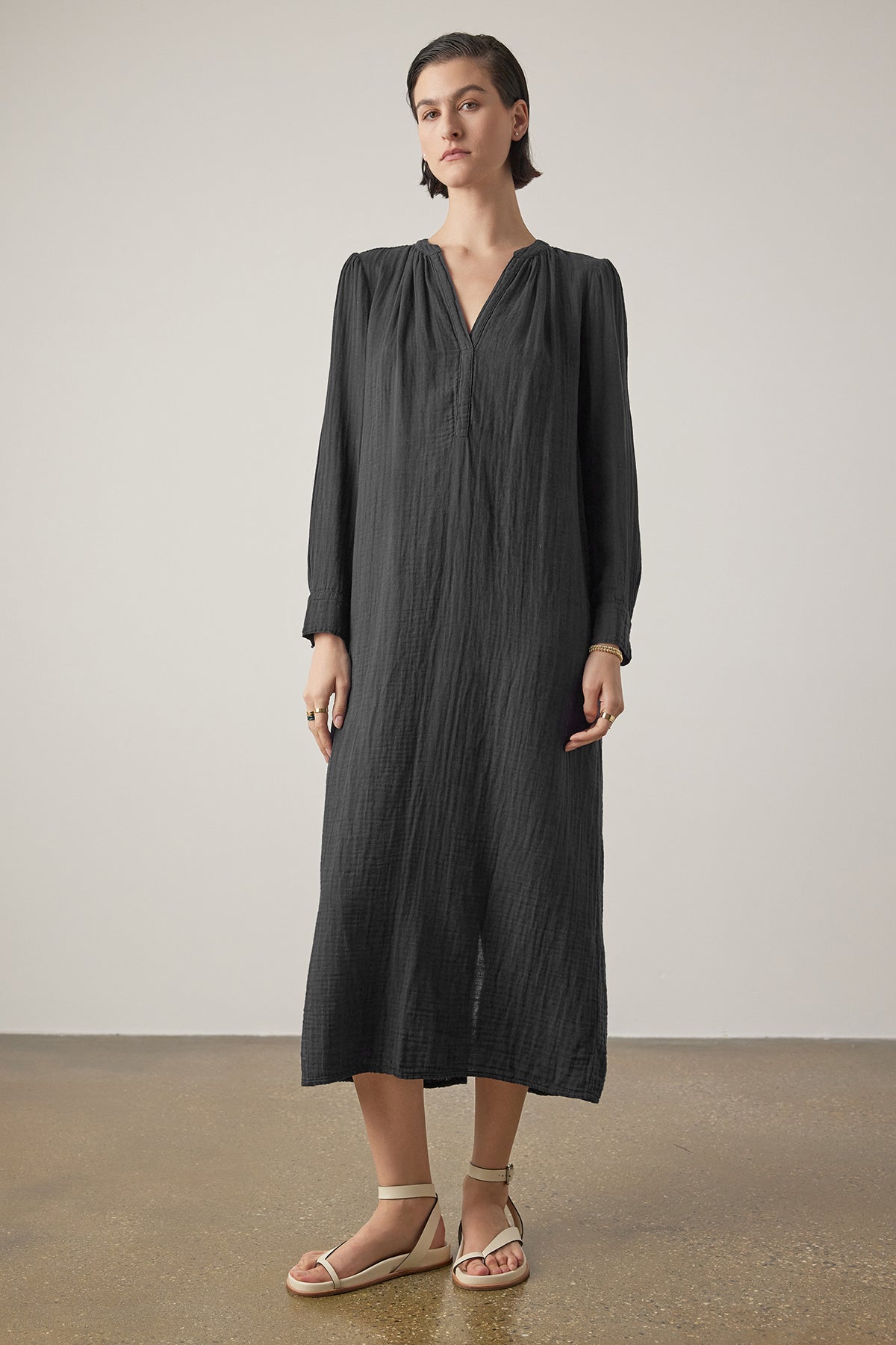 A person stands against a plain background, wearing a long-sleeved, ankle-length, dark gray DOHENY DRESS by Velvet by Jenny Graham with shirred seams and light-colored sandals. They have short hair and are looking directly at the camera.-36890839154881