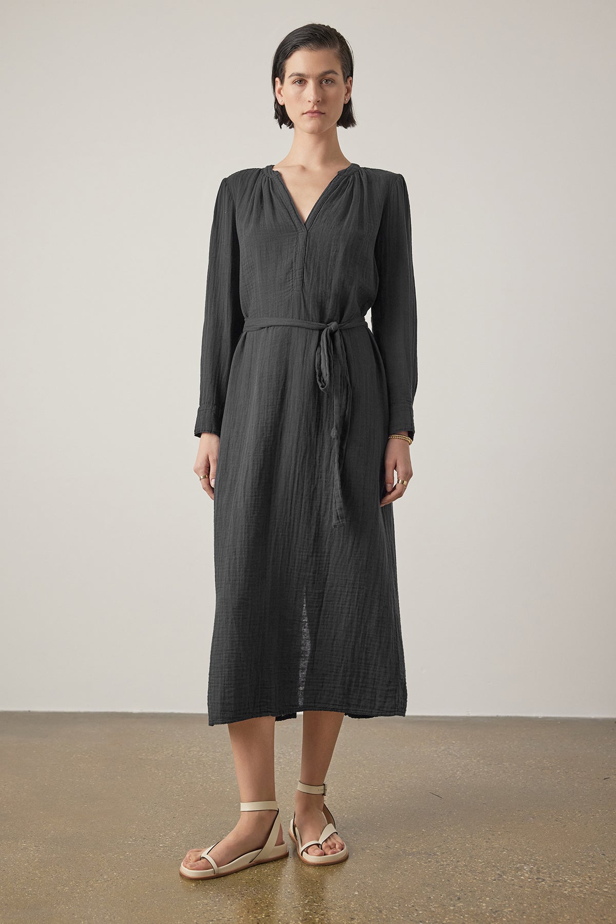   A woman stands against a plain background wearing the DOHENY DRESS, a long-sleeved, belted black dress with a V-neckline and shirred seams from Velvet by Jenny Graham. She is also wearing beige sandals. 