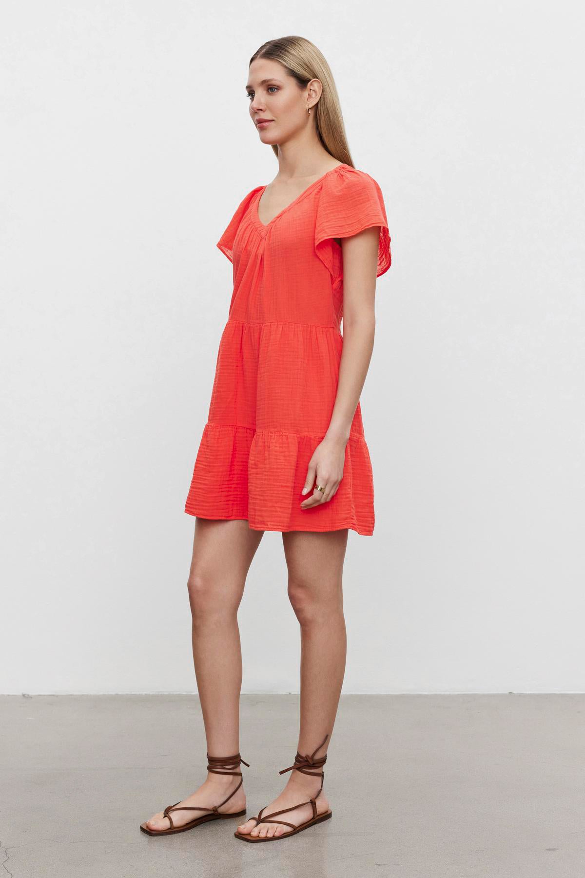 A woman stands against a plain background wearing a bright orange, Velvet by Graham & Spencer Eleanor Cotton Gauze Tiered Dress with tiered silhouette and brown strappy sandals.-36532828602561