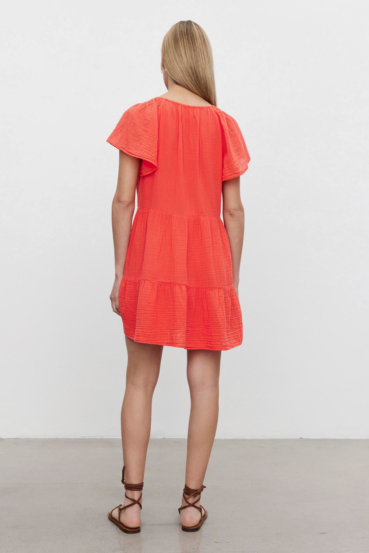 A woman from behind wearing a bright orange, short-sleeved Velvet by Graham & Spencer ELEANOR COTTON GAUZE TIERED DRESS, standing in a room with a white wall.-36532828635329