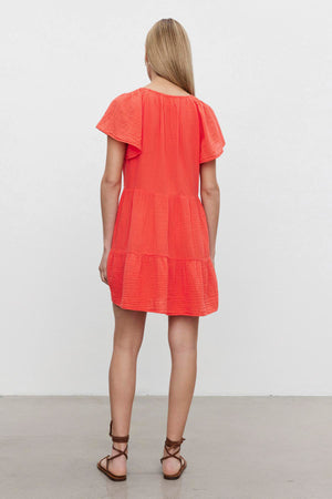 A woman from behind wearing a bright orange, short-sleeved Velvet by Graham & Spencer ELEANOR COTTON GAUZE TIERED DRESS, standing in a room with a white wall.