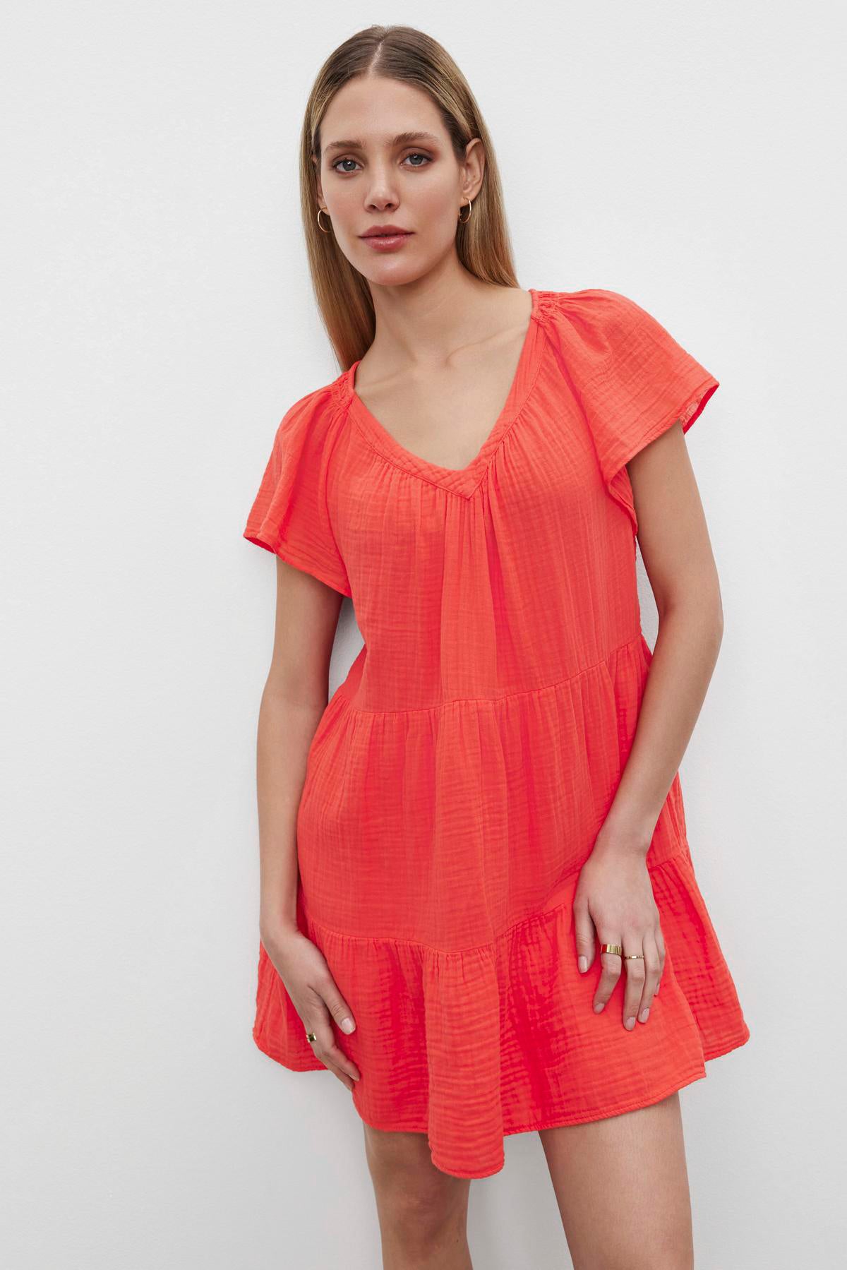 A woman in a bright orange, short ruffled Eleanor Cotton Gauze Tiered Dress by Velvet by Graham & Spencer standing against a white background, looking directly at the camera.-36532828569793