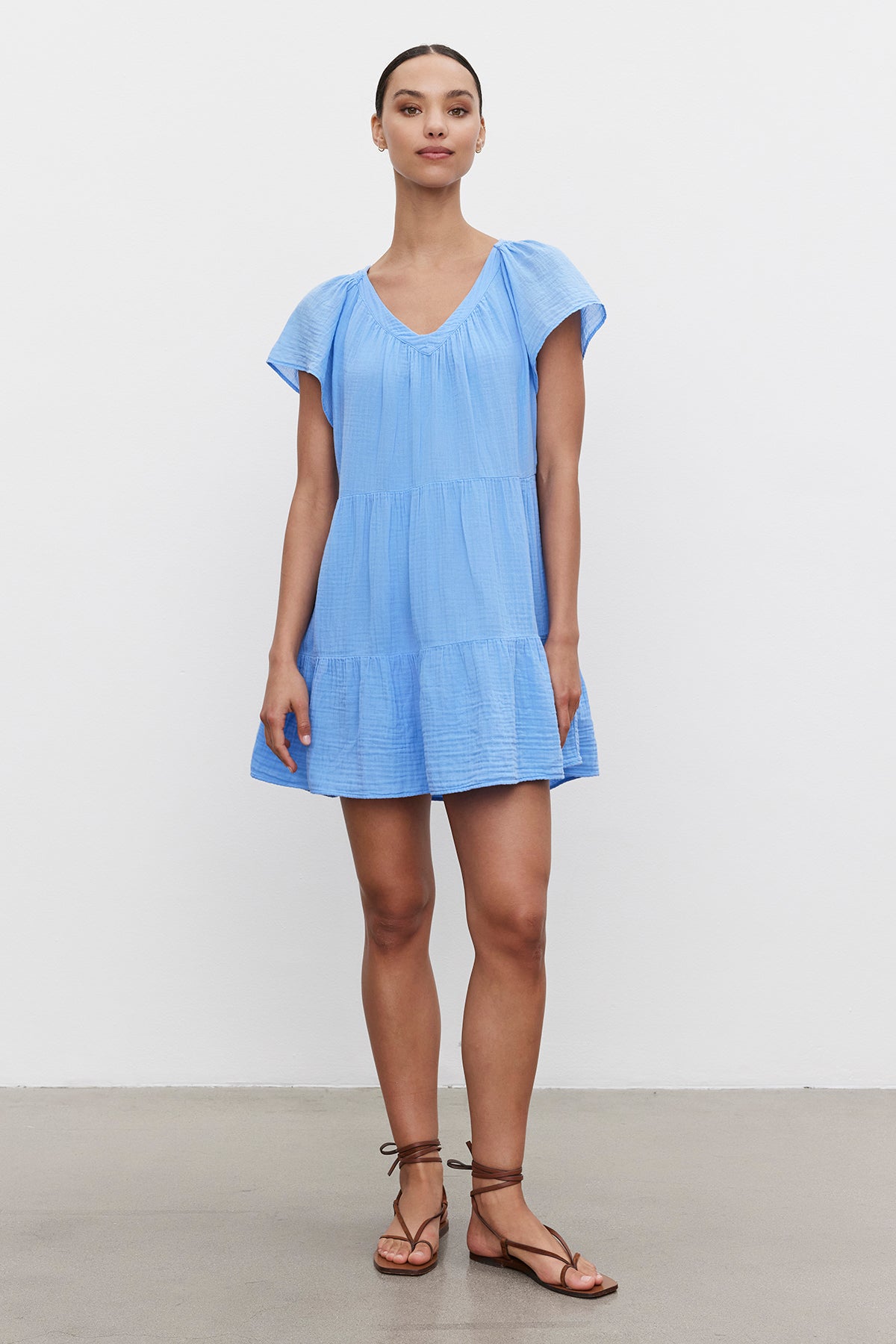 A woman in a light blue ELEANOR COTTON GAUZE TIERED DRESS by Velvet by Graham & Spencer with flutter sleeves and brown sandals stands confidently against a white background.-36443482620097