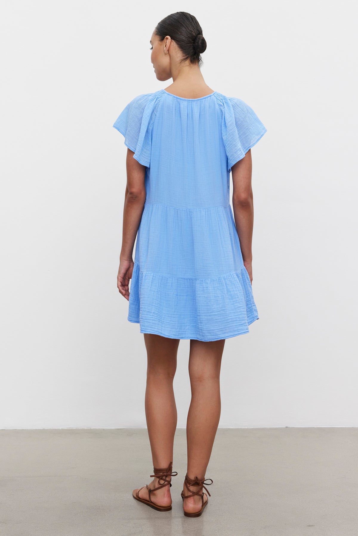   Back view of a woman standing in a gallery, wearing a light blue Eleanor Cotton Gauze Tiered Dress by Velvet by Graham & Spencer with flutter sleeves and brown strappy sandals. 