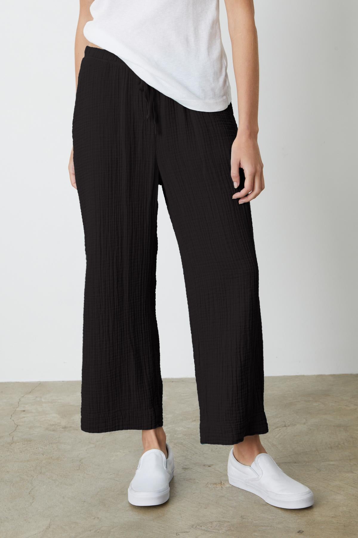   Person wearing black FRANNY COTTON GAUZE PANT by Velvet by Graham & Spencer, a white t-shirt, and white slip-on shoes, standing on a concrete floor against a plain white backdrop. 