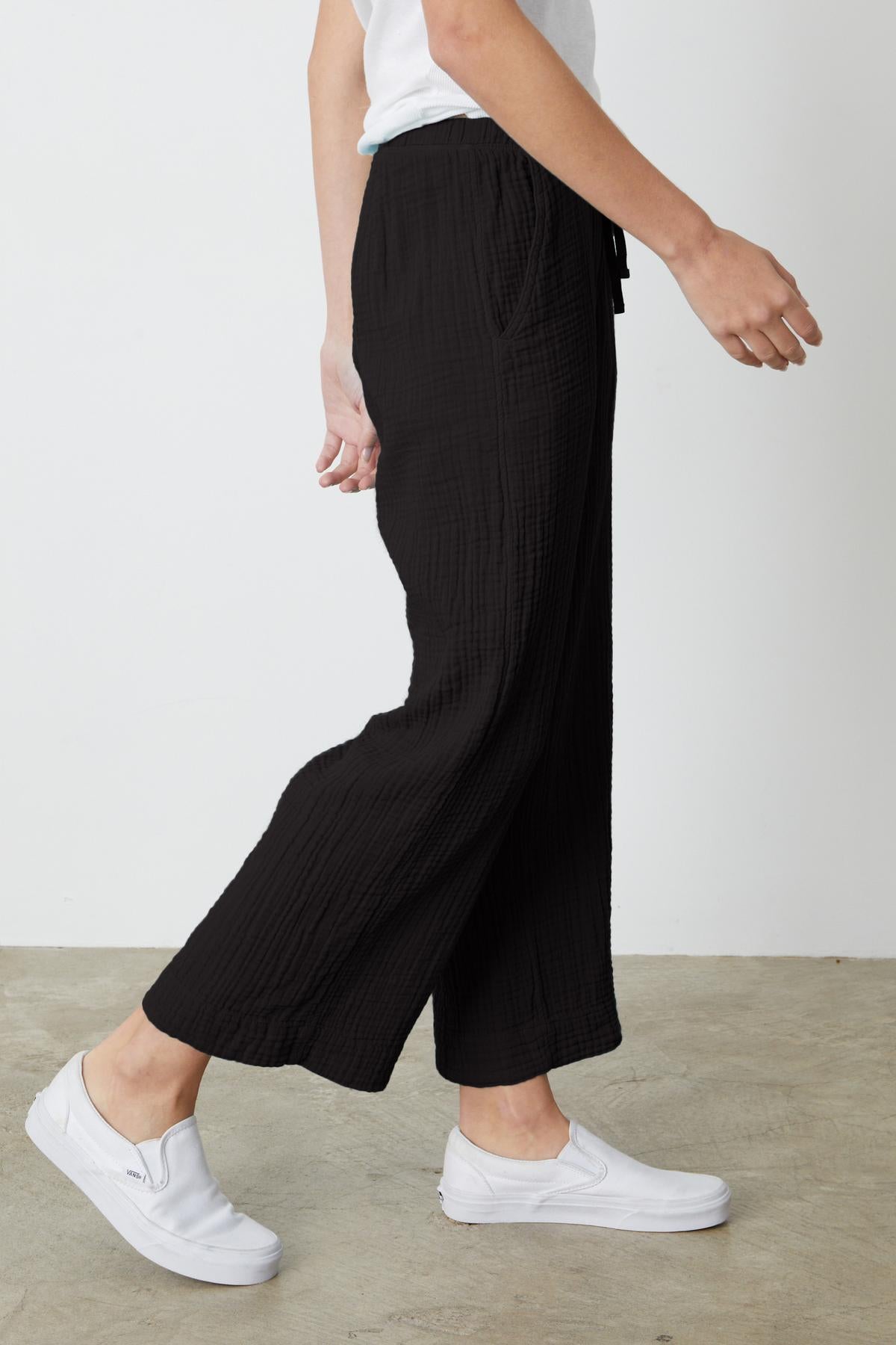 Person wearing black FRANNY COTTON GAUZE PANT by Velvet by Graham & Spencer with a relaxed leg and white slip-on shoes is shown from the side against a plain background.-37238901866689