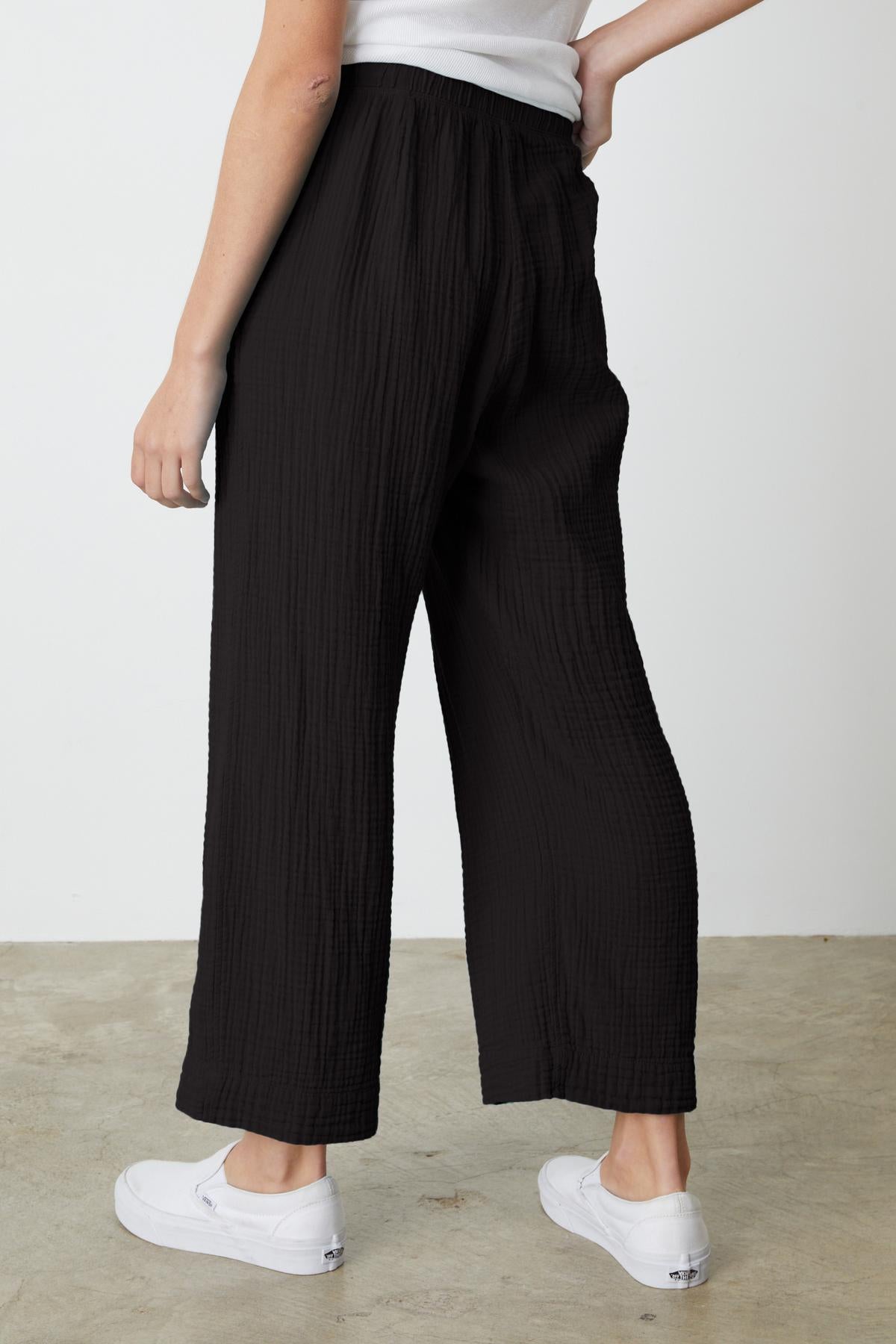 Person wearing black, loose-fitting FRANNY COTTON GAUZE PANT by Velvet by Graham & Spencer with a relaxed leg and a white top, standing sideways with one hand on hip. They are also wearing white sneakers.-37238901833921