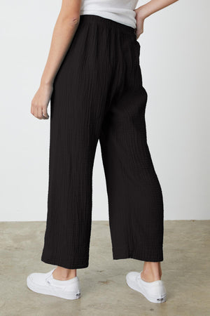 Person wearing black, loose-fitting FRANNY COTTON GAUZE PANT by Velvet by Graham & Spencer with a relaxed leg and a white top, standing sideways with one hand on hip. They are also wearing white sneakers.