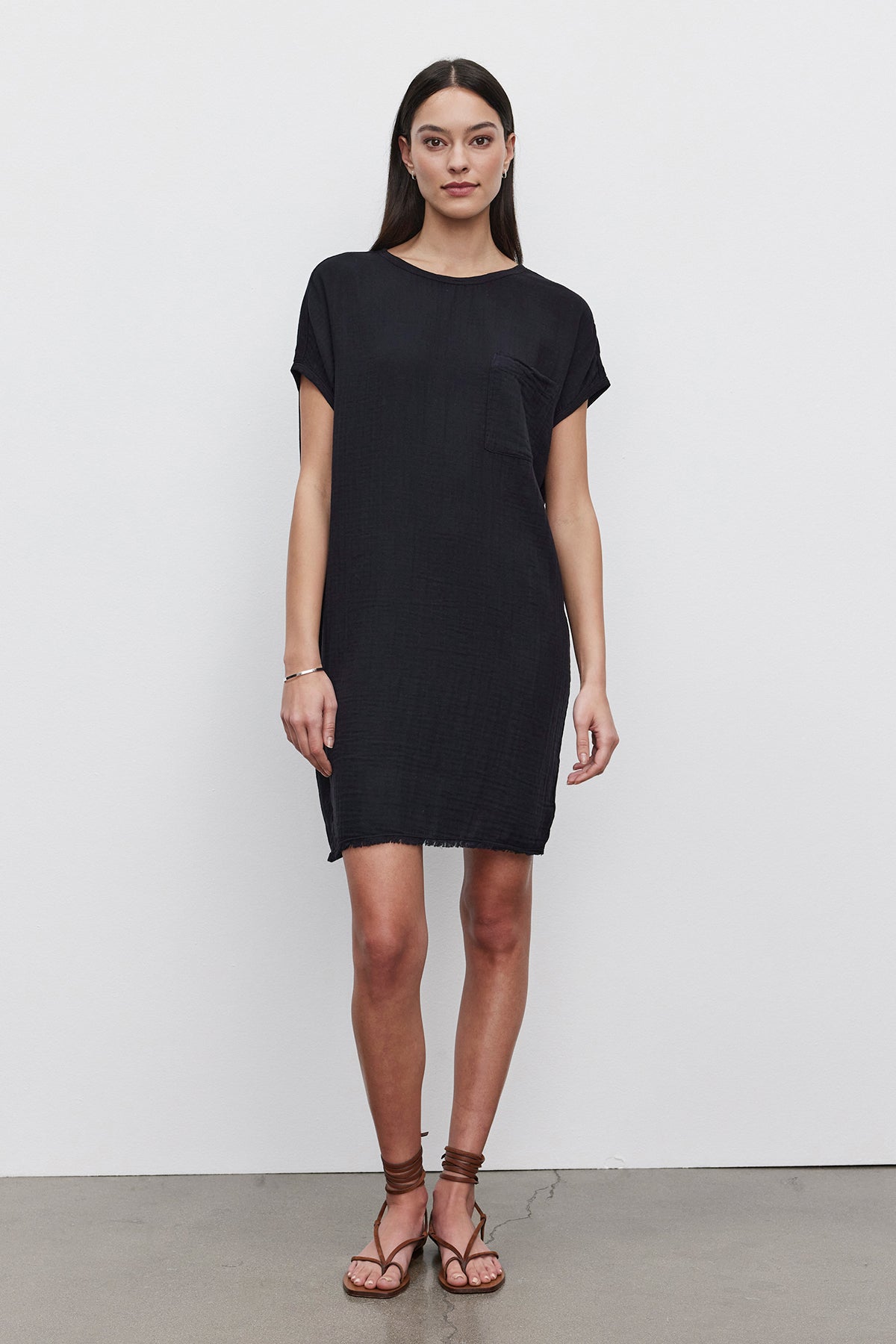 A woman in a short-sleeved, loose-fitting black HANNA DRESS by Velvet by Graham & Spencer with a small pocket on the left side stands against a plain white background. Made of soft cotton slub, her outfit pairs perfectly with brown sandals. She has long, straight hair cascading down to her shoulders.-36532922908865