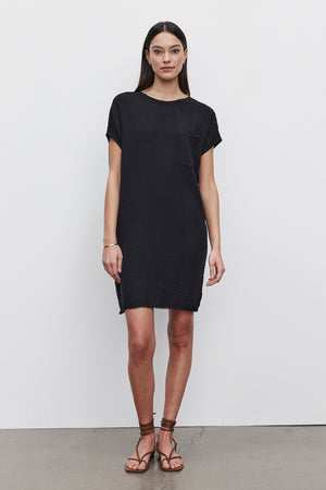 A woman in a short-sleeved, loose-fitting black HANNA DRESS by Velvet by Graham & Spencer with a small pocket on the left side stands against a plain white background. Made of soft cotton slub, her outfit pairs perfectly with brown sandals. She has long, straight hair cascading down to her shoulders.