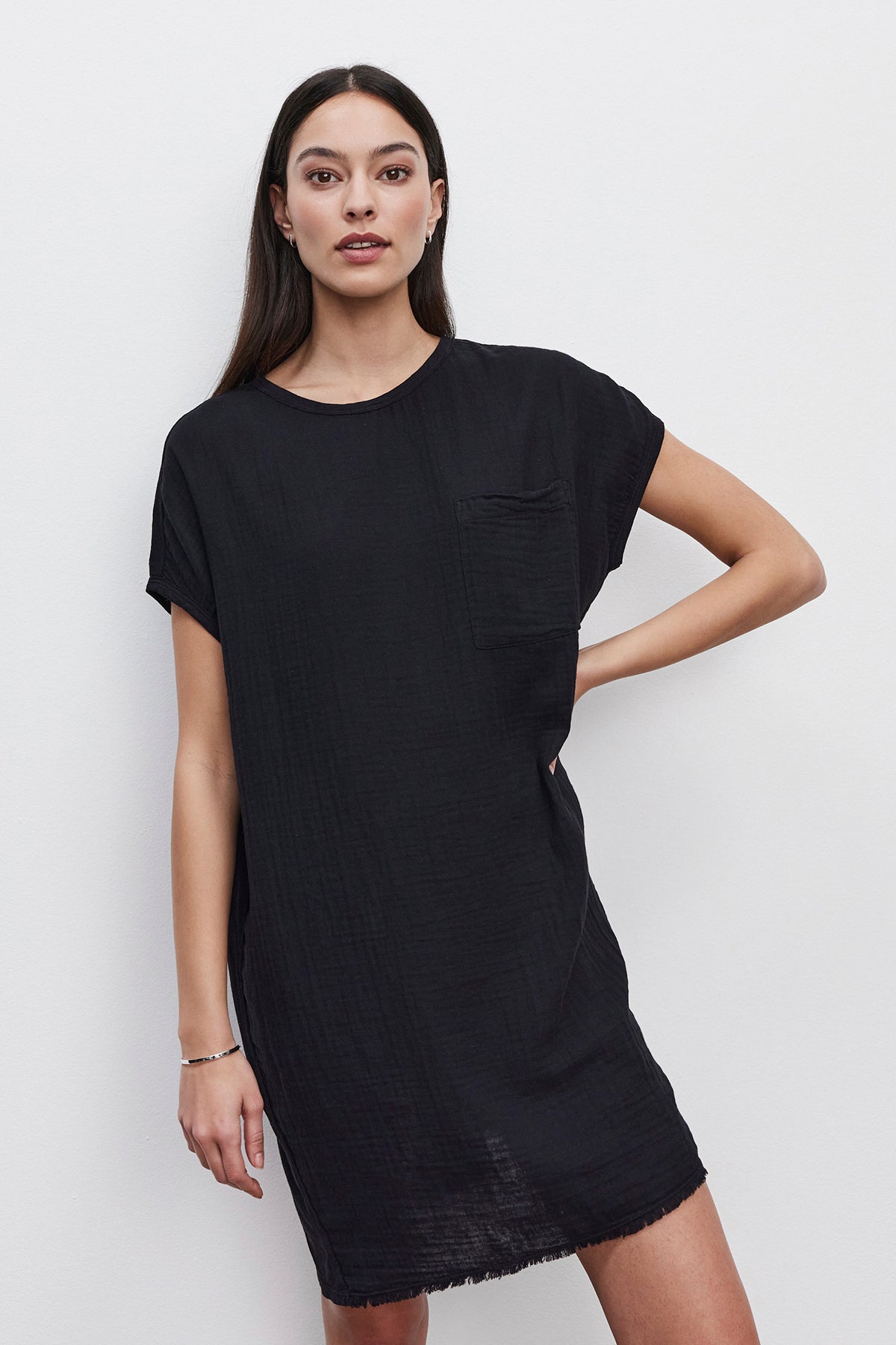 A person with long dark hair is wearing the HANNA DRESS by Velvet by Graham & Spencer, a simple black dress with short sleeves and a patch pocket made from cotton slub, standing against a plain white background.-36532922941633