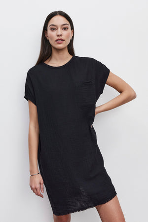 A person with long dark hair is wearing the HANNA DRESS by Velvet by Graham & Spencer, a simple black dress with short sleeves and a patch pocket made from cotton slub, standing against a plain white background.