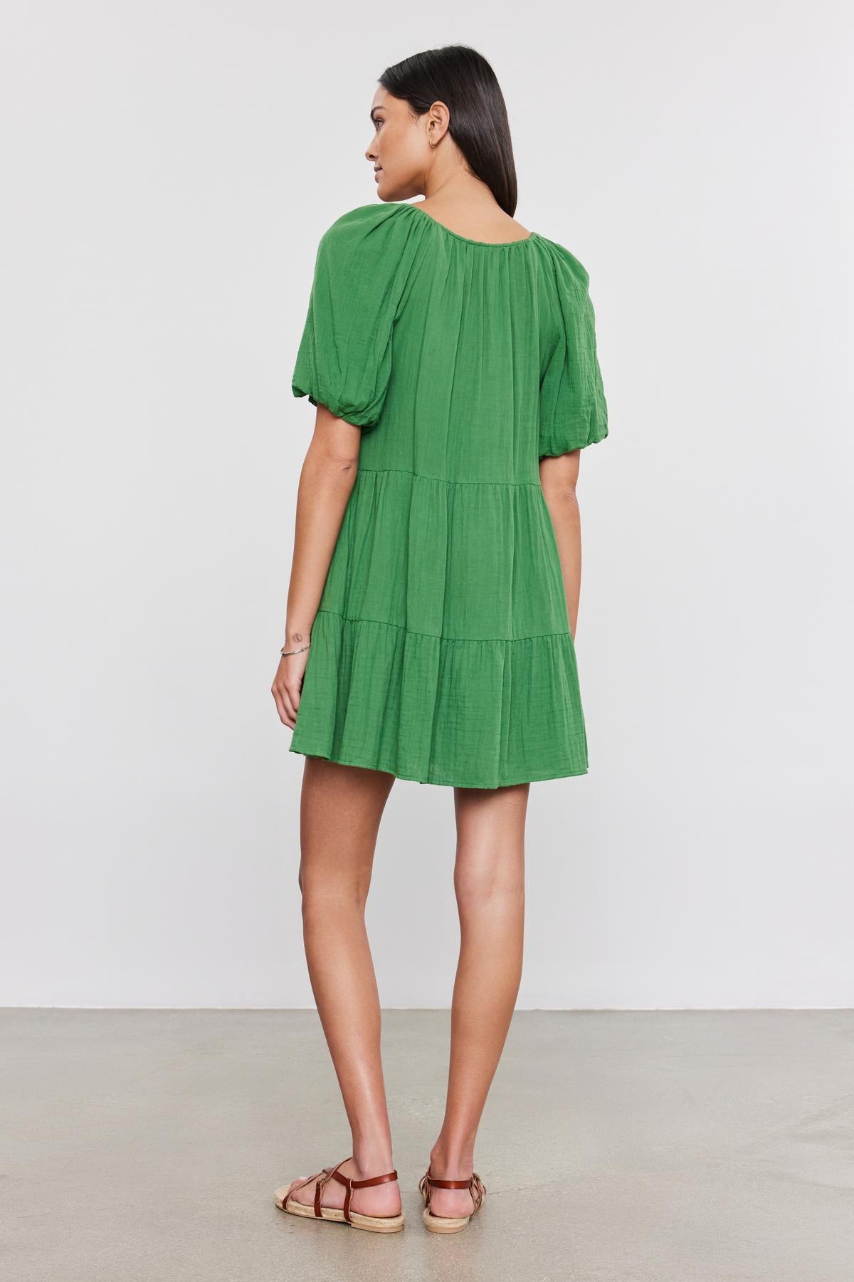   A woman wearing a green, two-tiered silhouette Velvet by Graham & Spencer HELENA COTTON GAUZE DRESS and brown sandals stands facing away, looking to her side against a neutral background. 