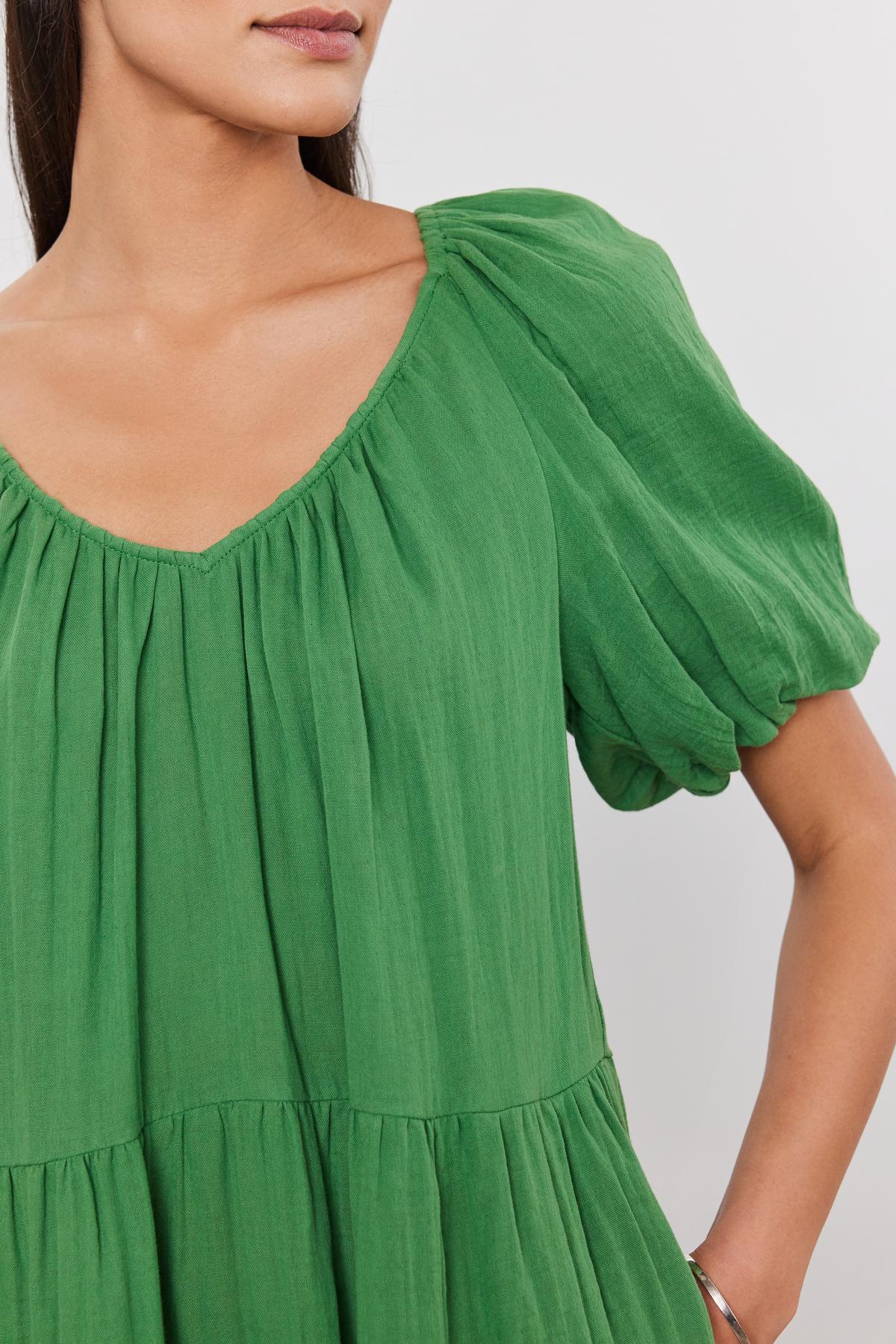   Close-up of a woman wearing a green Velvet by Graham & Spencer HELENA COTTON GAUZE DRESS with puff sleeves and a gathered neckline. Only her left shoulder and part of her face are visible. 