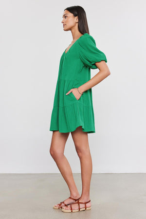 A woman stands in profile against a plain white background, wearing a HELENA COTTON GAUZE DRESS by Velvet by Graham & Spencer. The green dress features short puff sleeves, and she casually rests her hand in the pocket.