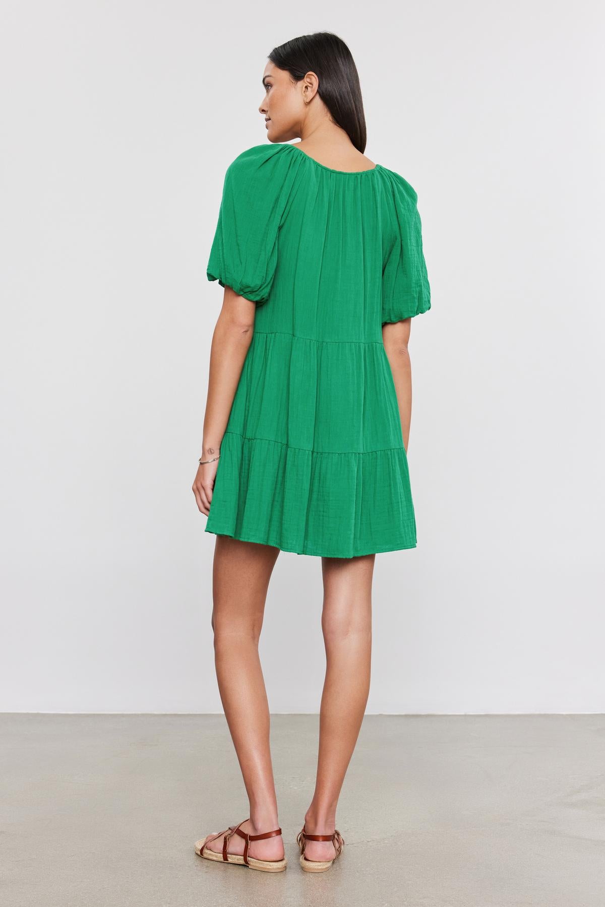   A person stands facing away from the camera, wearing the HELENA COTTON GAUZE DRESS by Velvet by Graham & Spencer with puff sleeves and sandals on a plain background. 