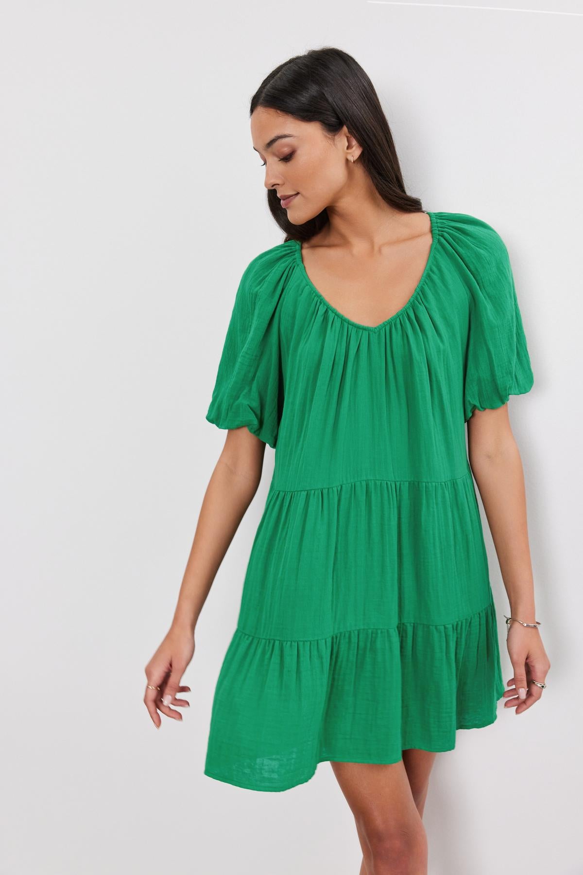   A woman in the Helena Cotton Gauze Dress by Velvet by Graham & Spencer, featuring a green hue, puff sleeves, and a V-neckline, stands against a plain white background. She is turned slightly to her right, looking downwards. 