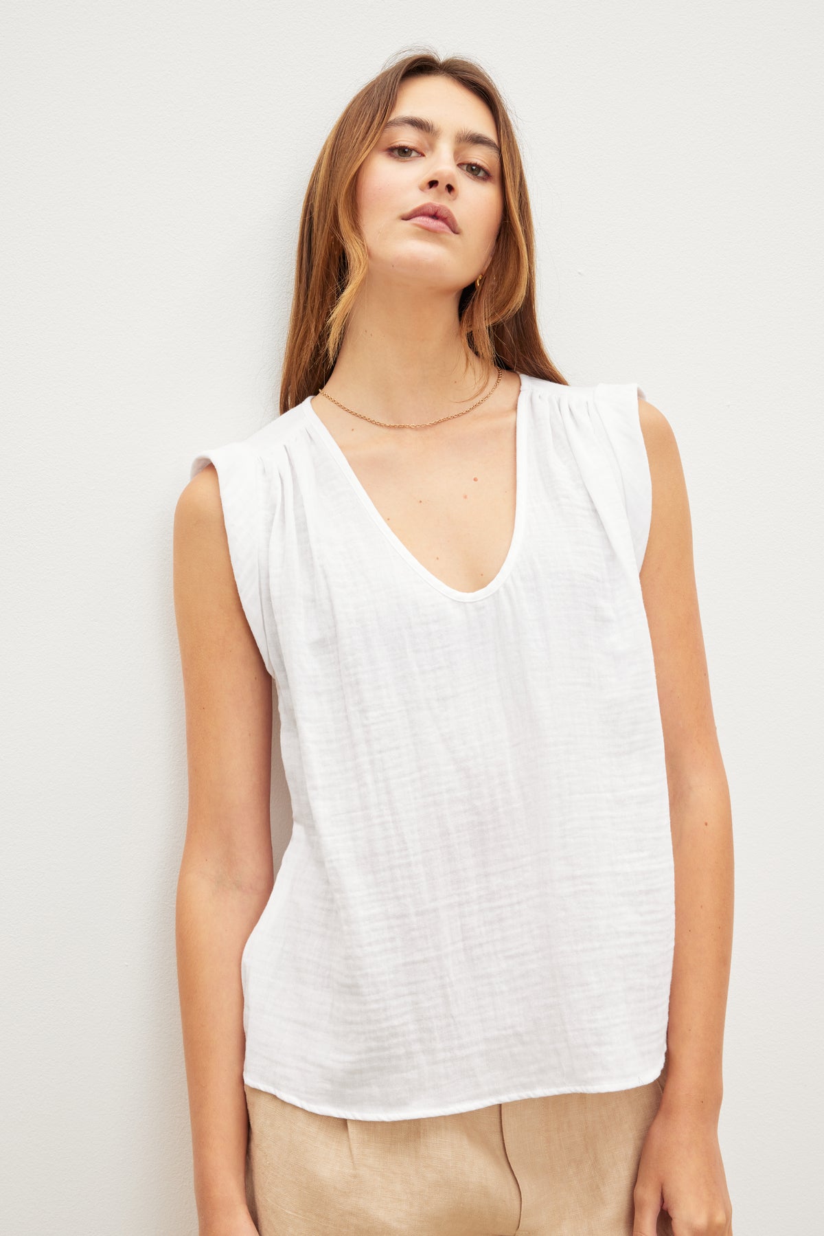 A person with long hair is wearing the Velvet by Graham & Spencer JAYLA COTTON GAUZE TANK TOP, a sleeveless white V-neck top with subtle shirred detailing, and beige pants, standing against a plain white background.-37618902302913