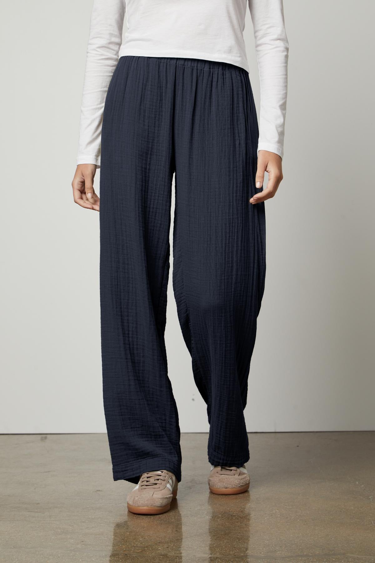   Woman standing in the Velvet by Graham & Spencer JERRY COTTON GAUZE PANT with an elastic waistband and white top, with hands slightly touching the pockets, paired with brown shoes. 