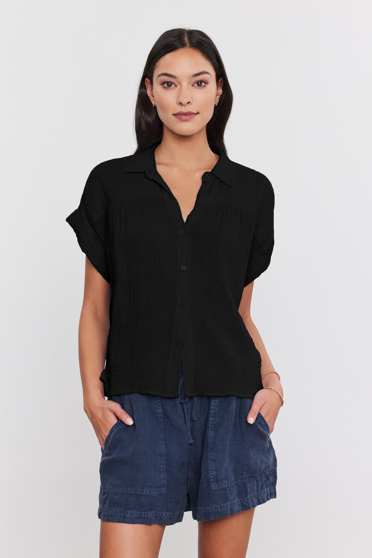 A woman stands facing the camera, wearing a JULIETA COTTON GAUZE SHIRT by Velvet by Graham & Spencer and blue shorts. She has a neutral expression and her hands are in her pockets.-37934927282369