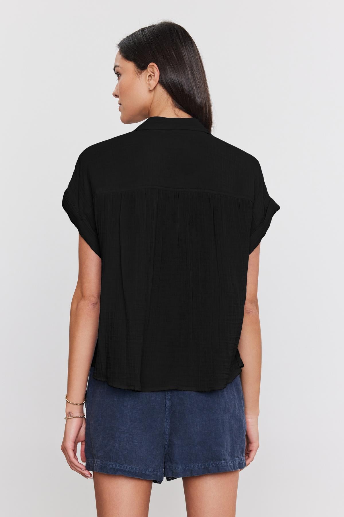 A woman with long dark hair is wearing a black short-sleeved Julieta Cotton Gauze Shirt by Velvet by Graham & Spencer and denim shorts, standing with her back to the camera.-37934927347905