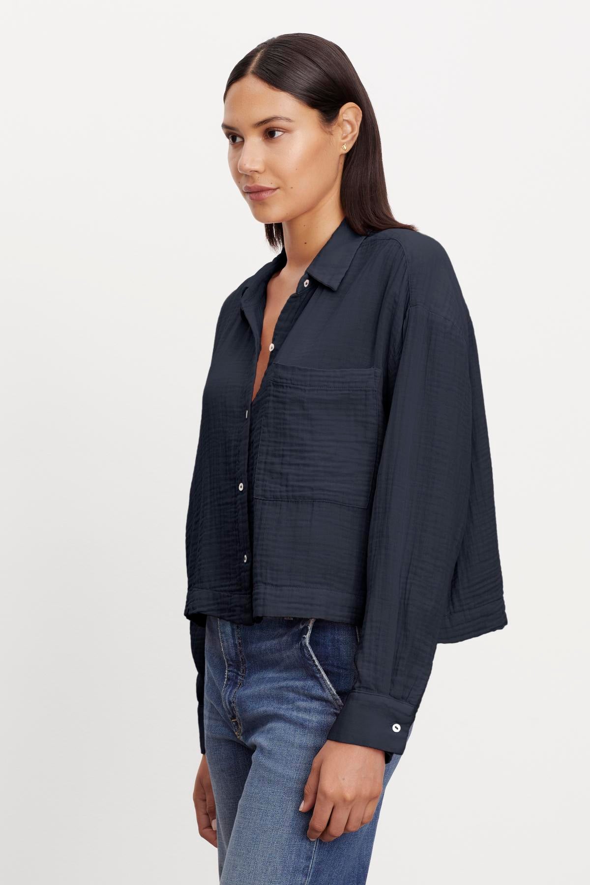   A woman wearing a dark Velvet by Graham & Spencer LANA COTTON GAUZE BUTTON-UP SHIRT and jeans stands in a side pose. 