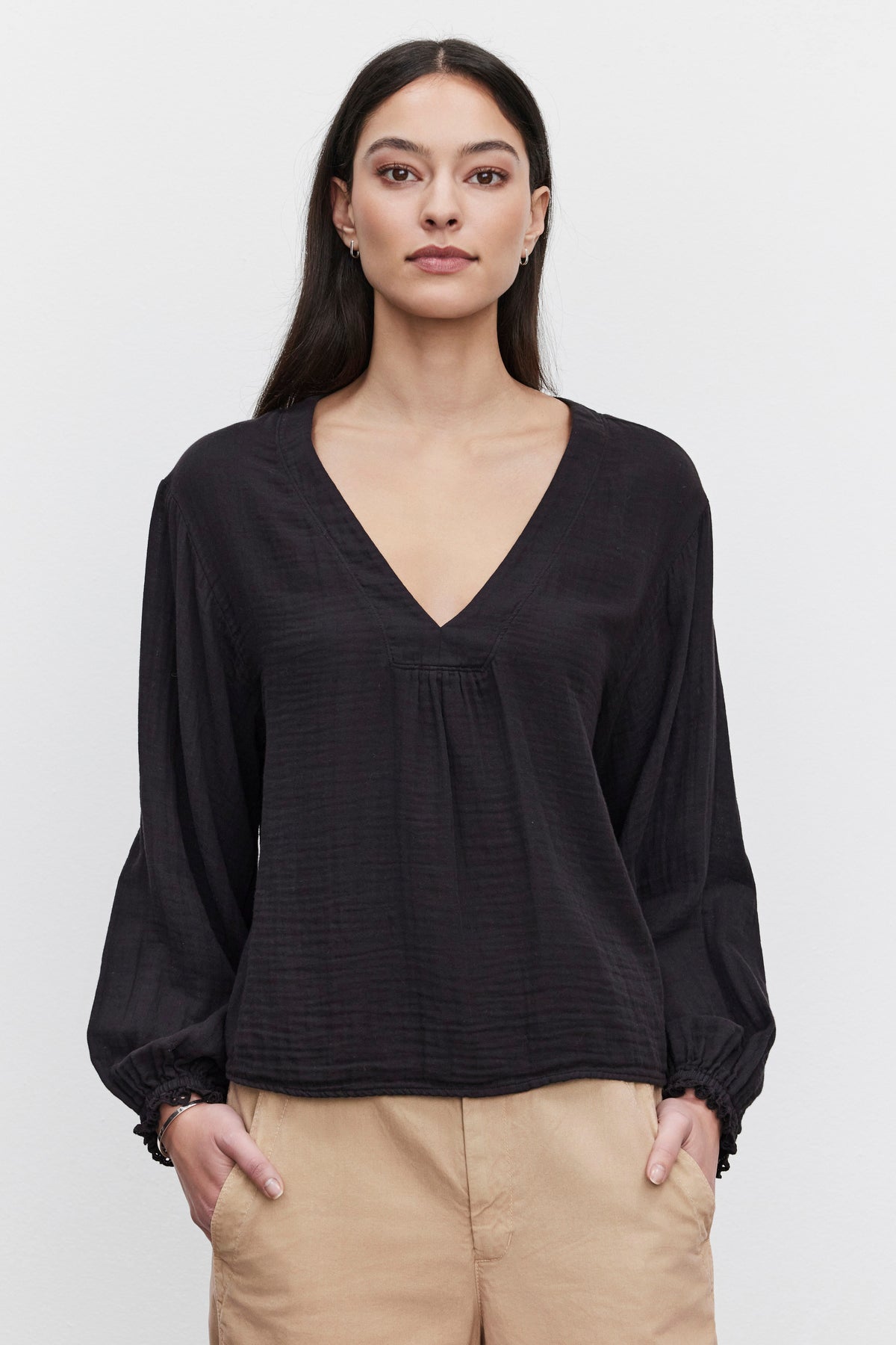   A woman with long dark hair stands against a plain white background, wearing a NAOMI COTTON GAUZE TOP by Velvet by Graham & Spencer. The black top features a V-neck and long sleeves, which she has paired perfectly with beige pants for a casual occasion. Her hands rest in her pockets as she gazes straight ahead. 