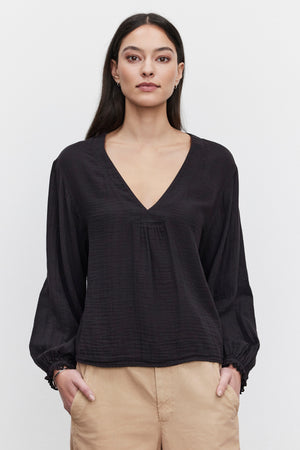 A woman with long dark hair stands against a plain white background, wearing a NAOMI COTTON GAUZE TOP by Velvet by Graham & Spencer. The black top features a V-neck and long sleeves, which she has paired perfectly with beige pants for a casual occasion. Her hands rest in her pockets as she gazes straight ahead.