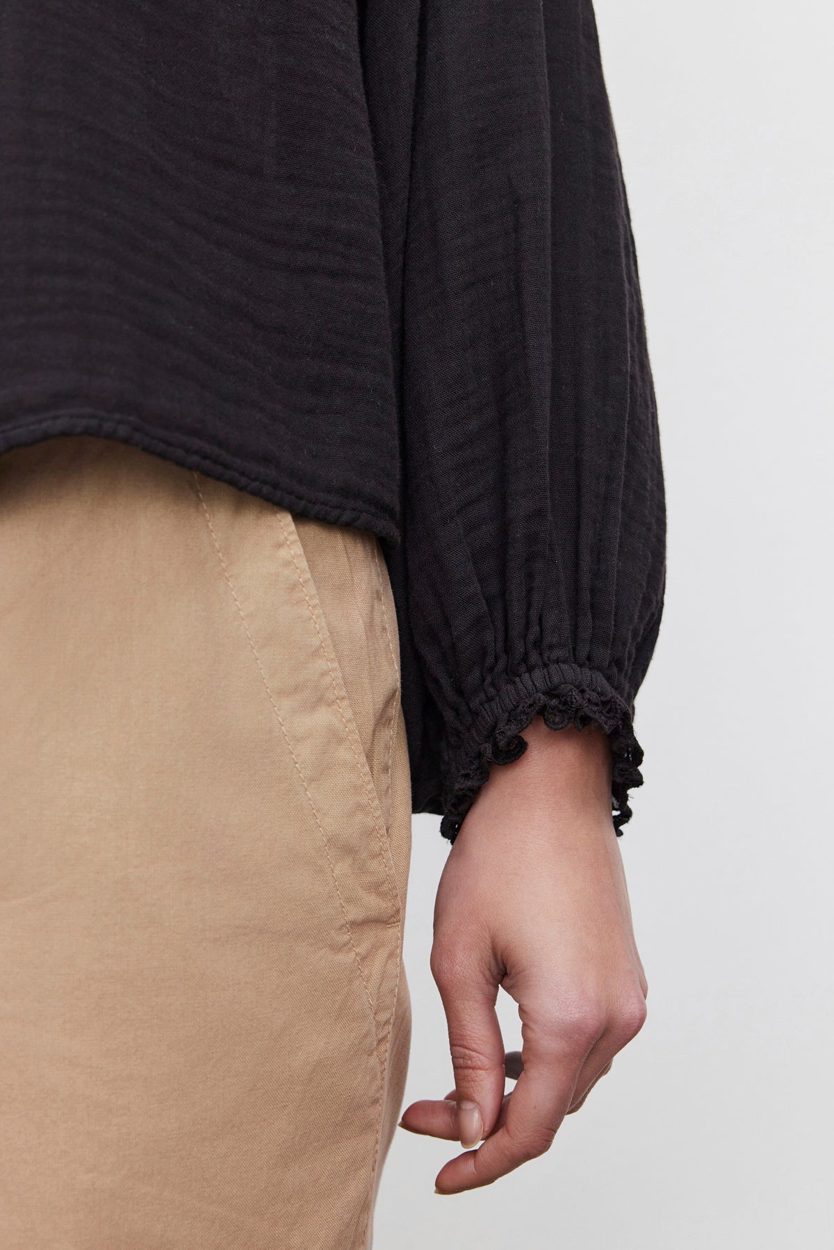   Close-up of a person wearing the NAOMI COTTON GAUZE TOP by Velvet by Graham & Spencer, a black long-sleeve shirt featuring textured fabric and airy cotton gauze, paired with beige pants. The hem of the relaxed-fit shirt ends at the waist, and the person's hand is relaxed at their side. 