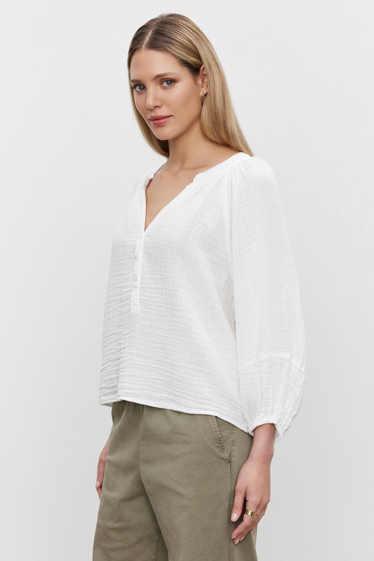   A woman with long blonde hair is wearing a VIVI COTTON GAUZE TOP, a relaxed-fit white long-sleeved blouse from Velvet by Graham & Spencer, paired with olive green pants. She is standing against a plain white background. 