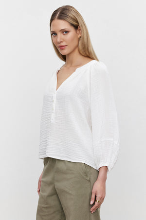 A woman with long blonde hair is wearing a VIVI COTTON GAUZE TOP, a relaxed-fit white long-sleeved blouse from Velvet by Graham & Spencer, paired with olive green pants. She is standing against a plain white background.