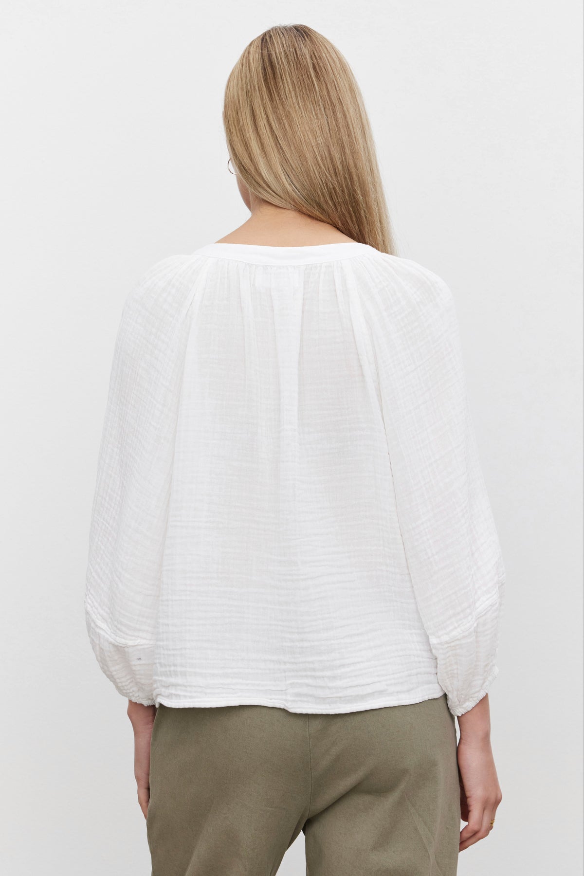   A person facing away, wearing the VIVI COTTON GAUZE TOP by Velvet by Graham & Spencer, a white long-sleeve textured blouse with a relaxed fit, paired with green pants, their long light brown hair cascading down. 