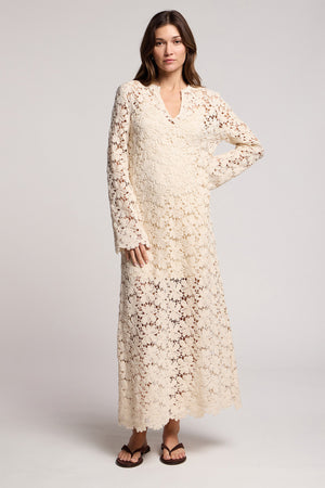 A woman stands against a plain background wearing the JOVIE DRESS by Velvet by Graham & Spencer, a cream long sleeve lace maxi dress with a V-neck, paired with brown sandals.